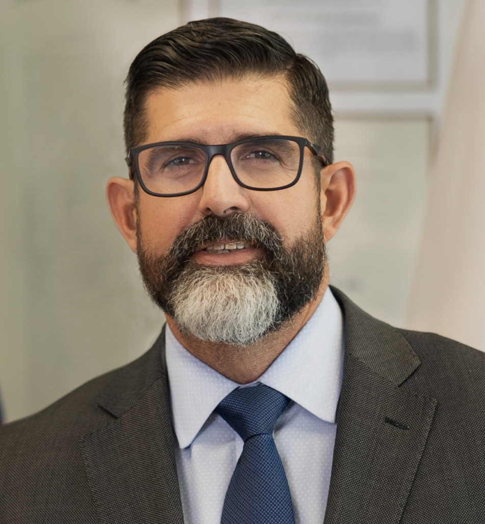 Academic freedom: Man with black hair and greying black beard and mustache on brown suit with blue shirt and tie stands between USA flag and Florida flag