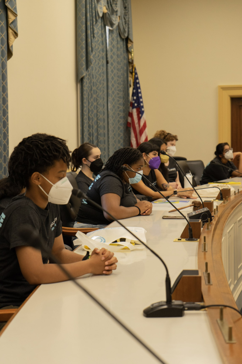 Youh Climate activists: Six young adults and teens sit with hands clasped at white table with microphones in front of each person.