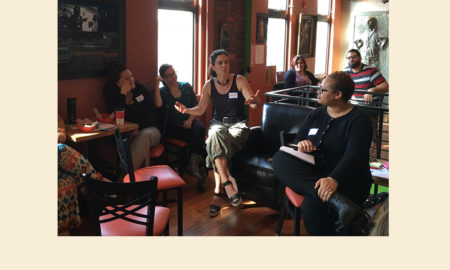 Youth development: 5 women sit in semicircle in restaurant talking, person speaking and gesturing has pulled-back dark hair, colorful necklace, black tank top, below-the-knee khaki pants and dark wedge sandals.