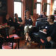 Youth development: 5 women sit in semicircle in restaurant talking, person speaking and gesturing has pulled-back dark hair, colorful necklace, black tank top, below-the-knee khaki pants and dark wedge sandals.