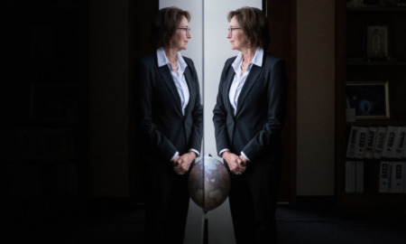 NM Foster Care: Barbara Vigil stands at window wearing dark suit and white blouse, looking at her reflection