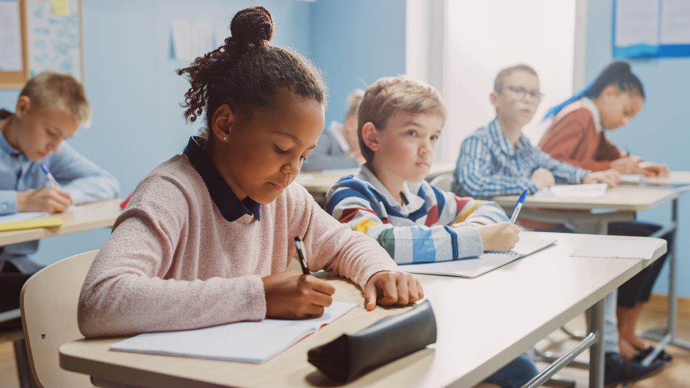 Pandemic School Funding: # elementary students cit in a row ar desks writing in papers while listening to teacher