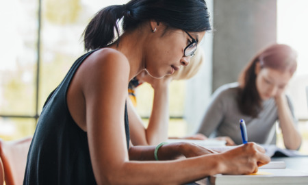 Close up shot of young woman writing notes with classmates studying in background.