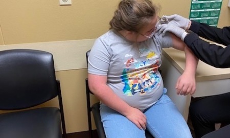 Down Syndrome vaccine effects: Young girl with blonde hair wearing glasses, light blue pants and light blue t-shirt, sits in a chair while someone wearing gloves and a dark clothes gives her a shot in upper left arm.