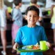 School meals report: happy child holding a tray of school food with other students lined up in background to get food