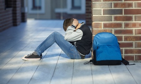 school violence prevention grants: young boy sitting against brick wall in hallway with hands on head and backpack on ground