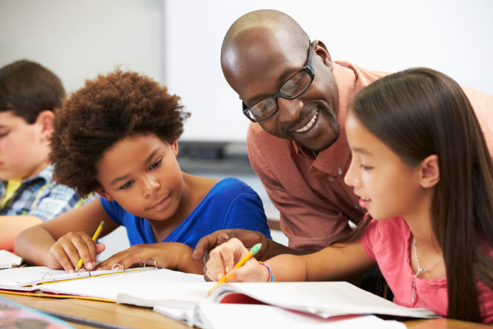 addressing student absenteeism, tutoring and afterschool programs: three pre-teen students sit working in notebooks while bald black man with dark glasses and red shirt explains lesson