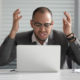 Grant makers hate 4 things: Black man with dark-rimmed glasses wearing gray suit and white shirt sits at silver laptop throwing hands in air with frustrated look