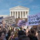 Nebraska abortion bans: Crowd of adults at rally with handmad signs held up gathered in front of multi-story, white building with 8 pillars in front