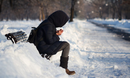 youth homelessness: lonely person with hoodie and backpack staying on bench in city park with snow