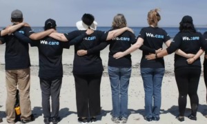 STEM education, hunger and community grants: people stand linking arms in chain on beach with shirts that say "we care"