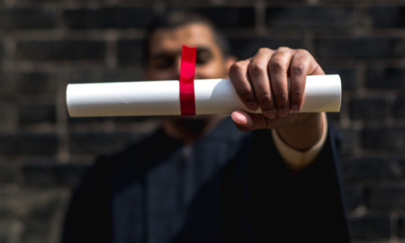 Prison college: Man burred in background wears college graduation gown holding rolled up white paper tied with red ribbon towards camera