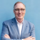 Tim Blaylock headshot: white man with dark-rimmed glasses in white shirt and blue blazer against light blue background