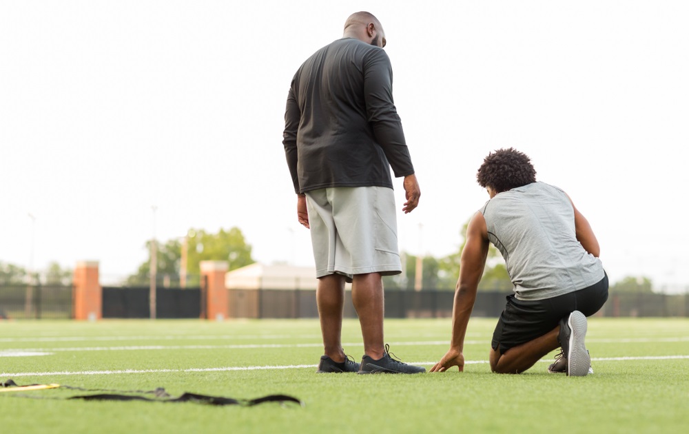 physical education: bald black man stands next to young black student kneeling on football field