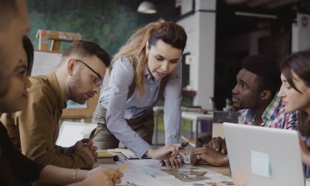 Youth leader development and career training grants: female team leader leans over table talking to team while pointing at things on table