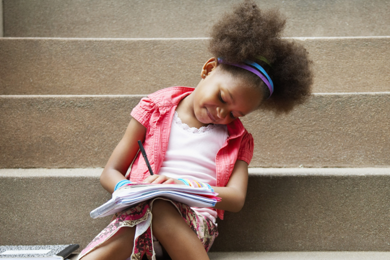 Language and Literacy: Young girl sitting on steps focused on reading and writing in an workbook on her lap.