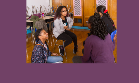 3 girls on chairs and 1 on the floor talk to each other in classroom with chairs up on desks