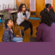 3 girls on chairs and 1 on the floor talk to each other in classroom with chairs up on desks