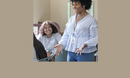 Florida: Beaming woman in flounced light blue top, jeans, necklace, earrings talks to three young women sitting down; one is turned to her and smiling.