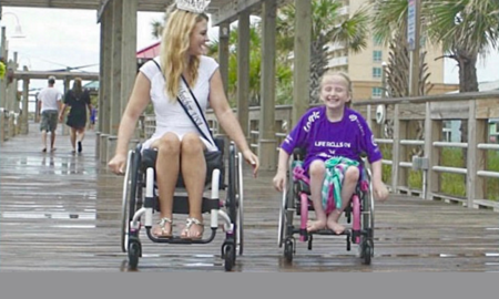 mentor: Blonde woman wearing tiara and white dress with diagonal sash in wheelchair laughs with little girl in purple T-shirt. Both are in wheelchairs on rain-soaked boardwalk.