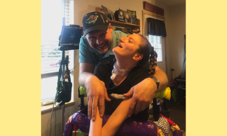 COVID vaccine access: Young woman with long blonde brais sit in wheelchair looking up at man in dark baseball cao standing behind her chair giving her a hug inside a home's living room