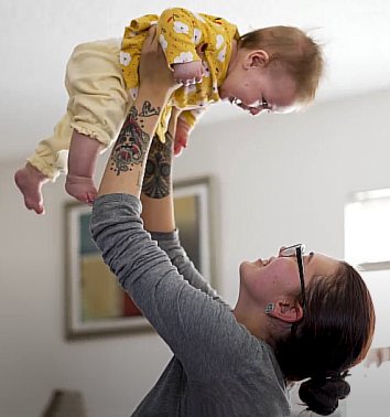 Homeles New Mexico youth; Homeless New Mexico youth:: Young woman with brown hair wearing glasses and a grey top holds a baby wearing yellow dress. up in the air.