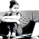 FAFSA and immigrant students: Ink drawing of young woman sitting on couch with arms wrapped around knees with sad, concerned expression on face while looking at laptop screen on low table on front of her